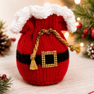 Puede incluir: Una bolsa de punto roja con cordón, borde superior de ganchillo blanco, una banda negra horizontal en el centro y una hebilla cuadrada de tono dorado. Un cordón metálico dorado con borlas sirve como cierre para la bolsa.
