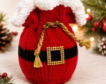 May include: A red knitted drawstring pouch featuring a white crocheted top trim, a horizontal black band around the center, and a square gold-toned buckle embellishment. A metallic gold cord with tassels serves as the closure for the bag.