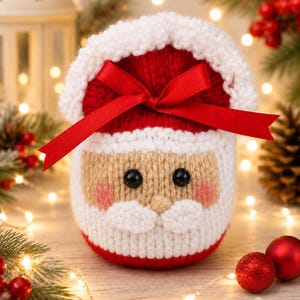 Può includere: Un sacchetto regalo cilindrico all'uncinetto a forma di Babbo Natale, con un cappello rosso e bianco dal bordo bianco, viso beige, occhi a perla neri, piccolo naso beige, baffi bianchi e guance rosa, chiuso in alto da un nastro di raso rosso.