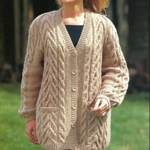 May include: A beige cable-knit cardigan with a V-neck, button closure, and two front pockets. The long-sleeved sweater has a textured design and is approximately 117cm long. The image also includes the text "aran" and size information.