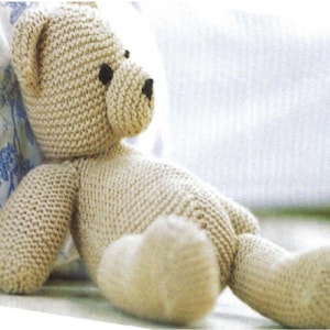 Könnte beinhalten: Handgefertigter, beigefarbener Strickteddybär mit schwarzen Knopfaugen und einer aufgestickten Nase. Der Bär sitzt und lehnt sich an ein Kissen mit floralem Muster. Die Arme und Beine des Bären sind sichtbar.