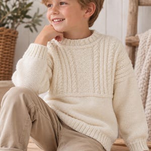 May include: A cream-coloured long-sleeve jumper featuring a ribbed crew neckline, cuffs, and hem. The garment displays a textured knit pattern on the sleeves and lower body, contrasted by vertical cable knit detailing across the upper chest area.