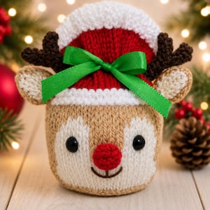 Puede incluir: Una funda de punto con forma de reno que presenta una cara beige y blanca con ojos de botón negros y una nariz roja. Lleva un gorro rojo y blanco adornado con astas de color marrón oscuro y un lazo de satén verde brillante al frente.