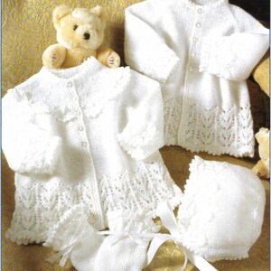 May include: A set of white knitted baby clothes, including a jacket, dress, hat, and mittens. The garments feature delicate lace detailing and are adorned with small buttons and satin ribbons. A teddy bear is nestled among the clothing.