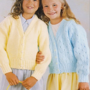 May include: Two young girls wearing knitted cardigans. One girl wears a pale yellow cardigan with pearl buttons, the other a light blue cardigan. Both cardigans have long sleeves. They are holding baskets with flowers.