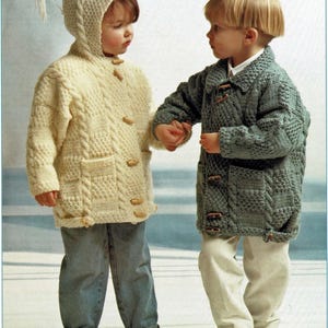 May include: Two children's knitted coats. One is cream-colored with a hood and wooden toggle buttons, paired with blue jeans. The other is a green-gray coat with wooden toggle buttons, paired with white corduroy pants. Both coats have pockets.