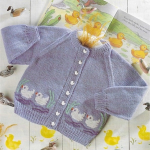 May include: A light purple knit cardigan featuring a button-down front with small white buttons. The lower portion of the garment displays an intarsia knit pattern of white ducks swimming in dark purple water with green reeds on both sides.