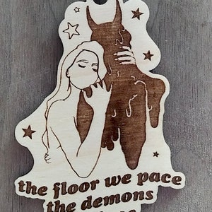 Könnte beinhalten: Holz-Anhänger mit einem Laser-geschnittenen Design einer Frau, die einen Dämon mit tropfenden Hörnern umarmt. Der Anhänger hat eine goldene Schnur zum Aufhängen und den Text "the floor we pace the demons we face".