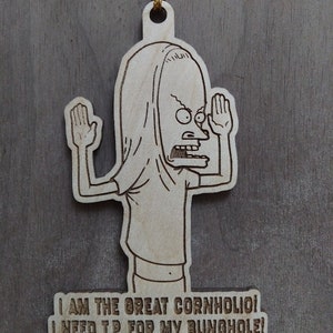 Op de afbeelding: Houten ornament met een stripfiguur met opgeheven armen en een schreeuwende uitdrukking. De tekst "I AM THE GREAT CORNHOLIO! I NEED T.P. FOR MY BUNGHOLE!" is gegraveerd. Een gouden draad is bevestigd om op te hangen.