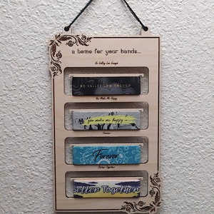 May include: Wooden wall-mounted organizer with five slots for displaying wristbands. The organizer has a decorative floral design and the text "a home for your bands..." at the top. The wristbands have various designs and text, including "No Valley Low Enough", "You Make Me Happy", "Forever", and "Better Together".