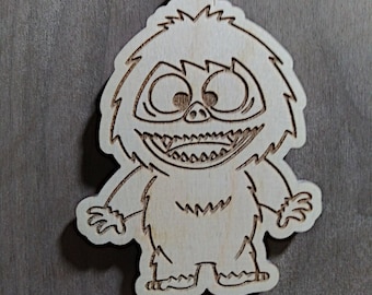 Puede incluir: Un adorno navideño de madera con forma de yeti de dibujos animados con una gran sonrisa y ojos grandes. El adorno es de color marrón claro y tiene un lazo de cuerda para colgar.