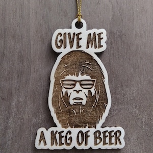 Puede incluir: Un adorno navideño de madera con una imagen de dibujos animados de una criatura con gafas de sol y el texto "Give me a keg of beer".