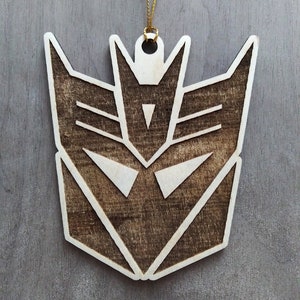 May include: A wooden Christmas ornament shaped like the Decepticons logo from the Transformers franchise. The ornament is brown with a white outline and is hanging from a gold string.