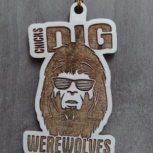 Puede incluir: Un adorno navideño de madera con un dibujo de un hombre lobo con gafas de sol y el texto "DIG CHICKS WEREWOLVES".