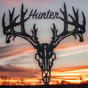 May include: Black metal deer skull wall art with the word "Hunter" above the antlers. The artwork is set against a sunset backdrop with a field in the foreground. A decorative piece for a hunting theme.