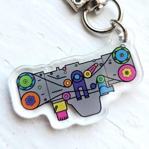 May include: A clear acrylic keyring with a colourful, abstract design of gears and machinery in shades of grey, purple, yellow, pink, blue, and green. The keyring has a silver clasp.