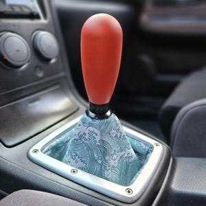 Peut inclure: Un pommeau de levier de vitesse rouge avec une base noire et un soufflet de levier de vitesse à motif de vagues vertes et blanches. Le soufflet porte du texte japonais.