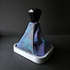 May include: A galaxy print gear shift knob with a black diamond-shaped top. The knob is mounted on a white base.