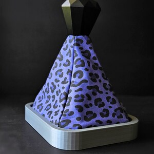 May include: A purple and black leopard print gear shift knob with a black diamond-shaped top. The knob is mounted on a gray base.