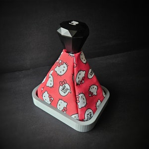 Peut inclure: Un pommeau de levier de vitesses blanc et gris avec un revêtement rouge à thème Hello Kitty. Le revêtement présente des personnages Hello Kitty et les mots "Hello Kitty".