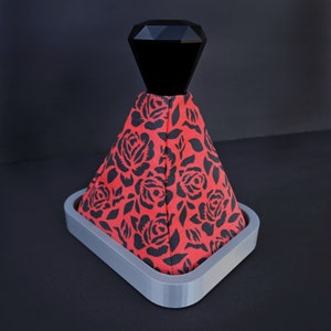 May include: A black and red gear shift knob cover with a rose pattern. The cover is made of fabric and has a black diamond-shaped top.