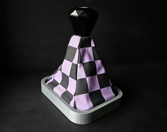 May include: A purple and black checkered gear shift knob with a black diamond-shaped top. The knob is mounted on a gray base.