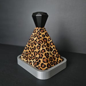 Peut inclure: Un pommeau de levier de vitesse décoratif avec un dessus géométrique noir et un cône en tissu à imprimé léopard. Le tissu est posé dans une base grise de forme carrée. Le fond est gris foncé uni.