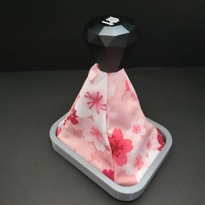 Peut inclure: Un soufflet de levier de vitesses à motif floral rose et blanc avec un pommeau de vitesse noir en forme de diamant. Le soufflet est fixé à une base grise.