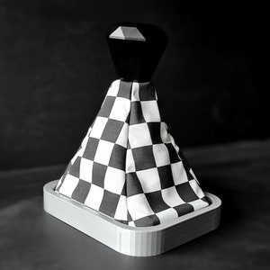 May include: A black and white chequered shift boot with a black top and a grey base. The shift boot is made of fabric and is designed to fit over a car's gear shift lever. The base is a rectangular shape.