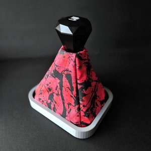 May include: A black diamond-shaped gear shift knob with a white logo sits atop a red and black patterned gear shift boot. The boot is attached to a white plastic base.