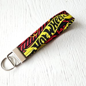 May include: A yellow, red and black fabric key ring with the words "Hot Wheels" printed on it. The key ring has a silver metal ring.