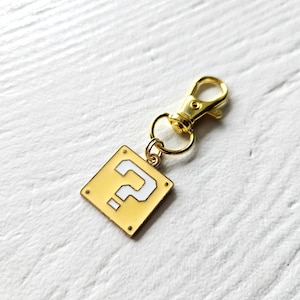 May include: Gold-coloured keychain with a question mark block charm. The charm is yellow with a white question mark and a gold border. The keychain has a gold-coloured lobster clasp.