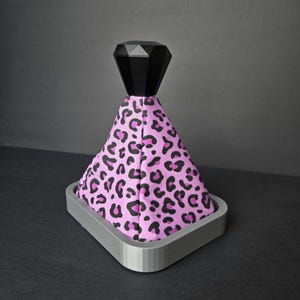 May include: A gear shift boot featuring a pink fabric with a black leopard print pattern. The top of the boot is capped with a black, faceted, diamond-shaped knob. The base is secured within a rectangular, light gray, 3D-printed plastic frame.