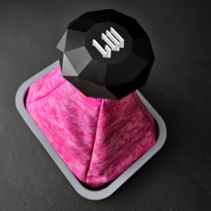 Könnte beinhalten: Ein schwarzer, diamantförmiger Schaltknauf mit den Buchstaben "LW" oben, der auf einem rosafarbenen und weißen, gemusterten Schaltknaufbezug sitzt.