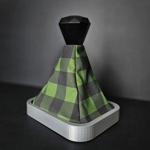 May include: A car gear shift boot with a black geometric top and a green and gray plaid fabric cover. The boot sits in a silver-colored rectangular base, creating a modern, stylish look. The background is a dark gray.