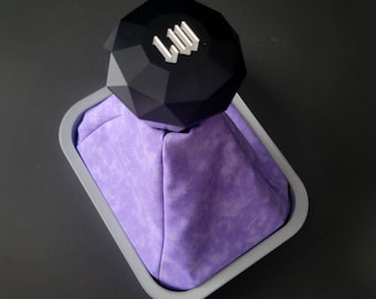May include: A black, faceted object with the letters "LW" in silver sits atop a purple fabric-lined, gray rectangular container. The container is set against a dark gray background.