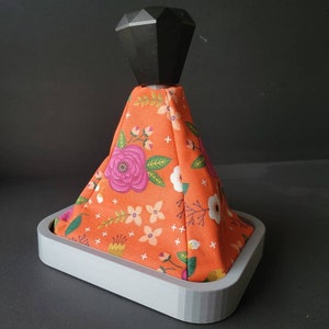 May include: A shift boot with a floral pattern in orange, pink, and white. The boot is topped with a black, geometric shift knob and sits in a gray base. The fabric features a repeating pattern of flowers and leaves.