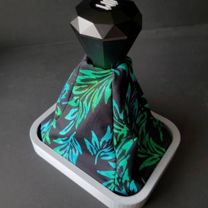 May include: A black diamond-shaped gear shift knob with a white base sits on a white base with a black and green patterned fabric cover.