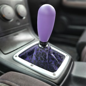 May include: A purple shift knob with a black base and a floral pattern shift boot. The shift knob is a solid, smooth color. The shift boot has a black rose pattern on a purple background. The shift knob is in a car interior.