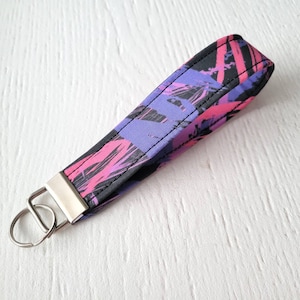 May include: A black key chain with a pink and purple abstract paint splatter design. The key chain has a silver metal key ring.