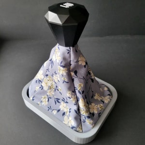 May include: A grey floral patterned gear shift knob with a black diamond-shaped top. The knob is sitting on a grey base.