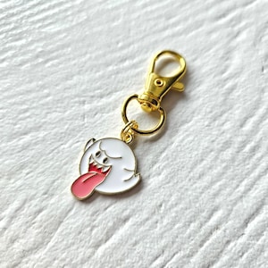 May include: Gold keychain featuring a white ghost with a red tongue sticking out. The ghost has black eyes and a toothy grin.