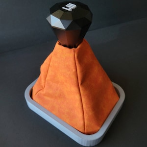 Peut inclure: Un pommeau de levier de vitesses en forme de diamant avec une finition noire et une base grise. Le pommeau est monté sur une base grise avec un revêtement en tissu orange.