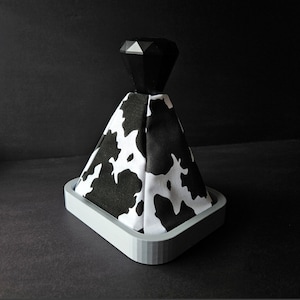 Peut inclure: Un pommeau de levier de vitesses en imprimé vache noir et blanc avec un dessus en forme de diamant noir. Le pommeau est monté sur une base grise.