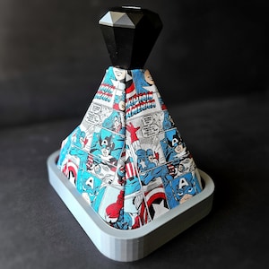 May include: A grey, rectangular base with a pyramid-shaped top covered in a comic book print featuring Captain America. The top of the pyramid has a black, faceted, diamond-shaped piece.
