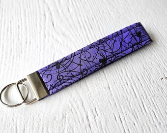 May include: Purple fabric key chain with a black spider web and spider design. The key chain has a silver metal key ring.