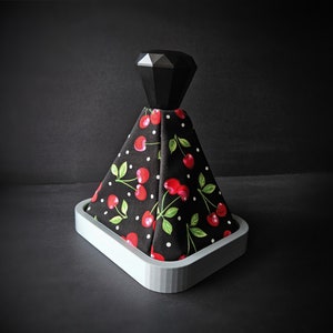 Peut inclure: Un couvre-pommeau de levier de vitesse de voiture en tissu imprimé cerises noir et blanc avec un dessus en forme de diamant noir. Le couvre-pommeau est sur une base grise.