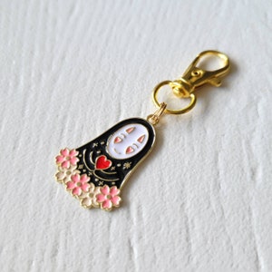 May include: Gold keychain featuring a black and white No Face character from Spirited Away with pink cherry blossoms and a red heart.