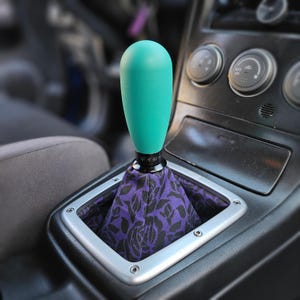 May include: A teal gear shift knob with a black base and a purple floral patterned boot. The text "シフト" is printed on the base of the knob.