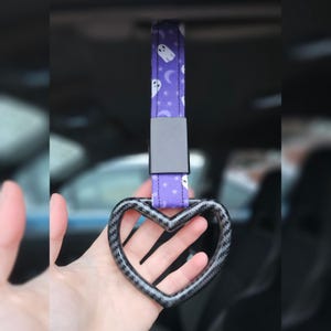 May include: A purple car handle strap with a heart-shaped grip. The strap features a pattern of white ghosts, moons, and stars. The grip has a carbon fiber design and is connected to the strap with a black metal piece.